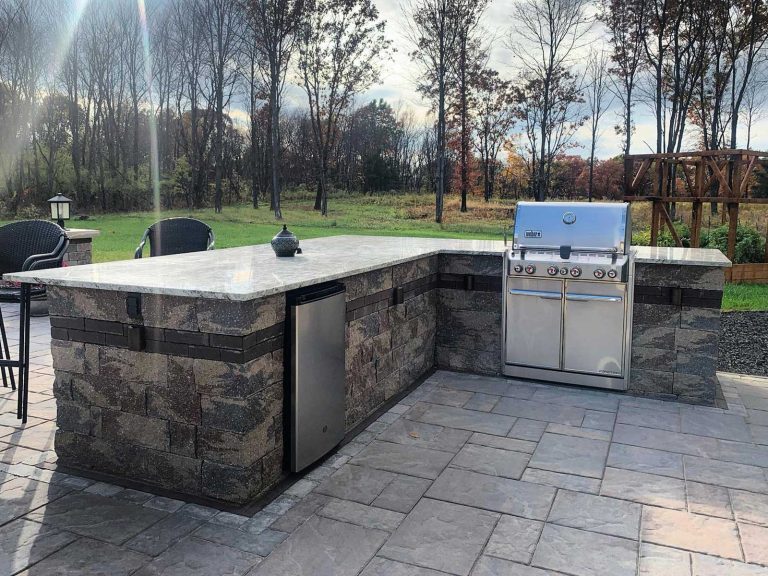L-shaped outdoor kitchen made with Unilock block, surrounding a paver patio in a backyard in Clifton Park, NY